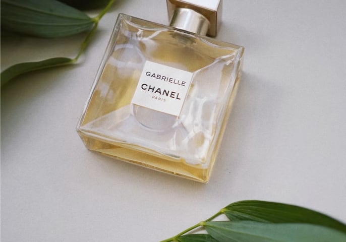 A bottle of Gabrielle Essence Eau De Parfum lays on a white surface between two leafy stems